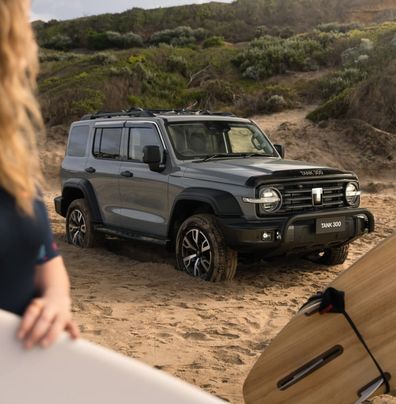 Fossil Grey GWM Tank 300 with a black nudge bar parked on the sand with people walking by carrying surfboards