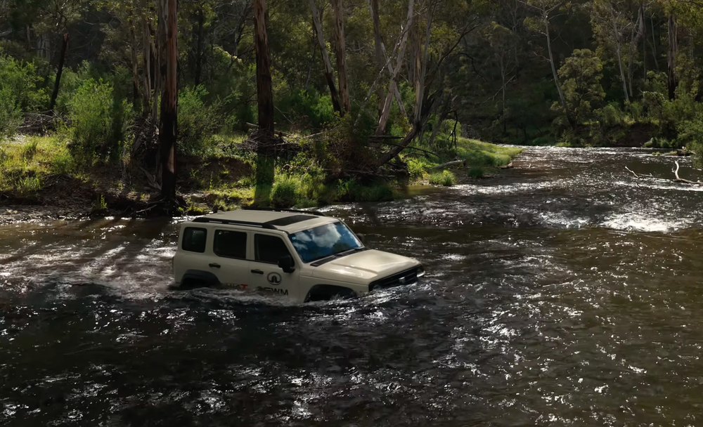 A GWM Tank 300 SUV crossing a shallow, flowing river in a lush forest setting under dappled sunlight.