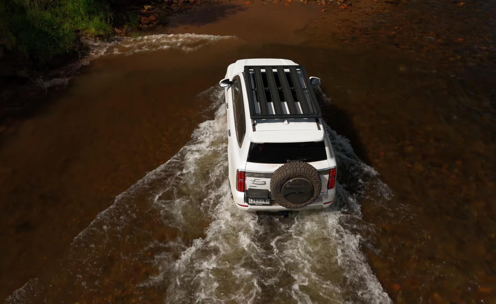 White SUV with a roof rack and spare tire navigating through a shallow river, surrounded by rocks and greenery.