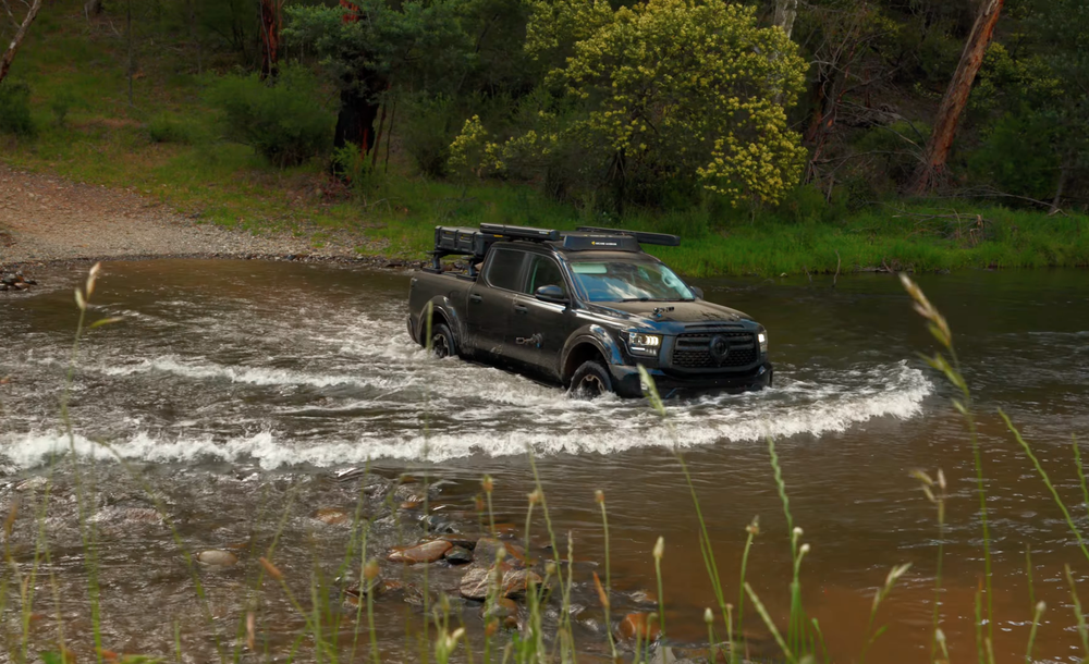 A black off-road truck drives through a shallow river in a lush, green forest setting.