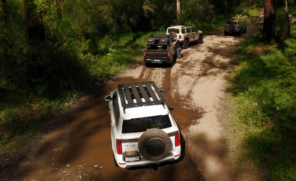 Four off-road vehicles driving on a muddy forest trail surrounded by lush green trees, with one person standing beside the second car.