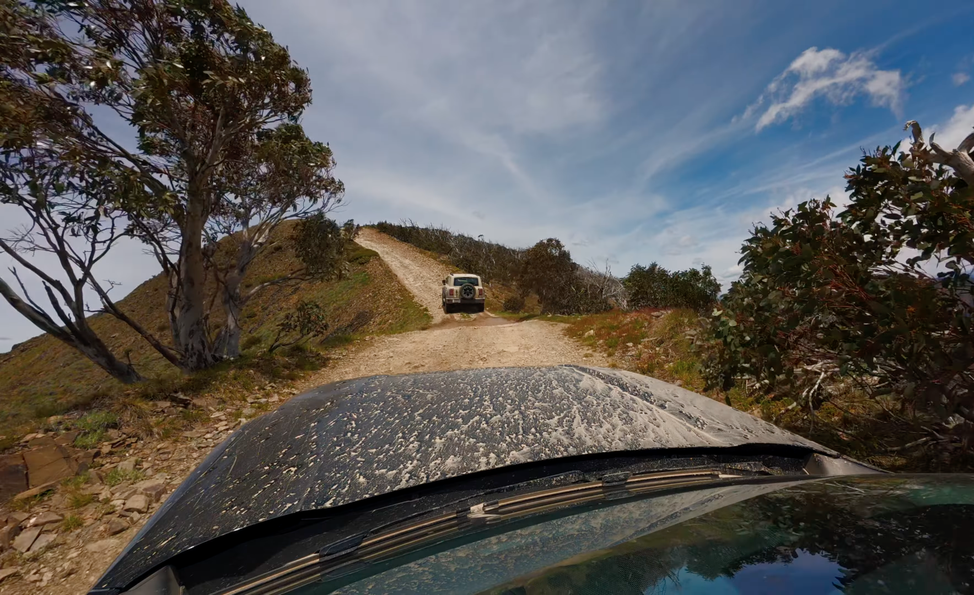 A white off-road vehicle drives up a dirt trail on a hillside, surrounded by trees under a partly cloudy sky.