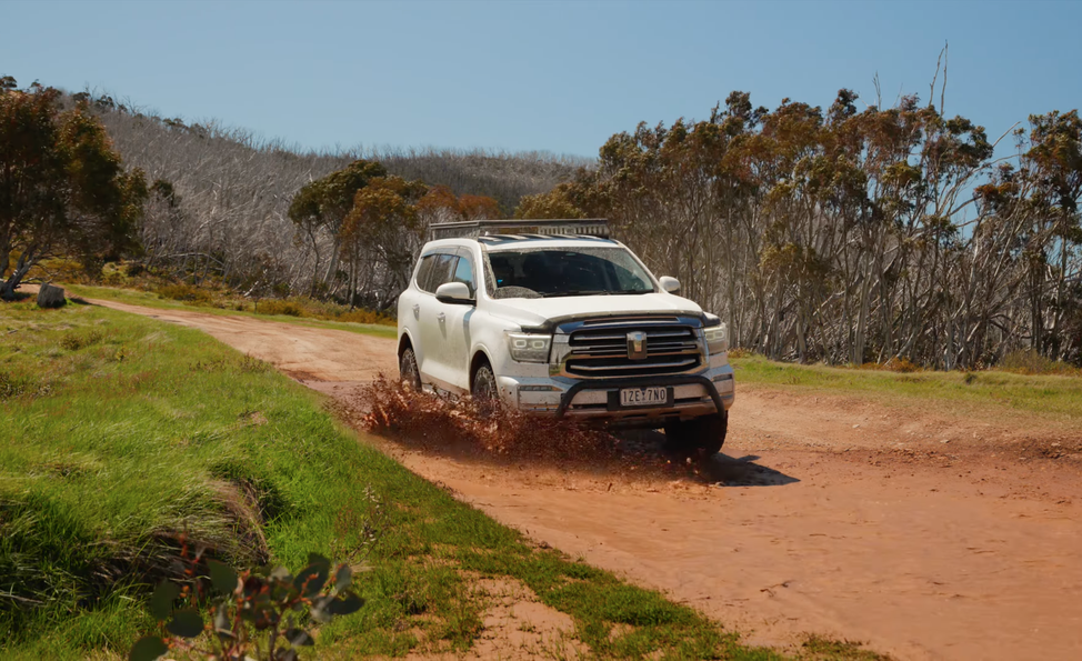 White SUV driving through a muddy puddle on a dirt road, surrounded by trees and green grass under a clear blue sky.