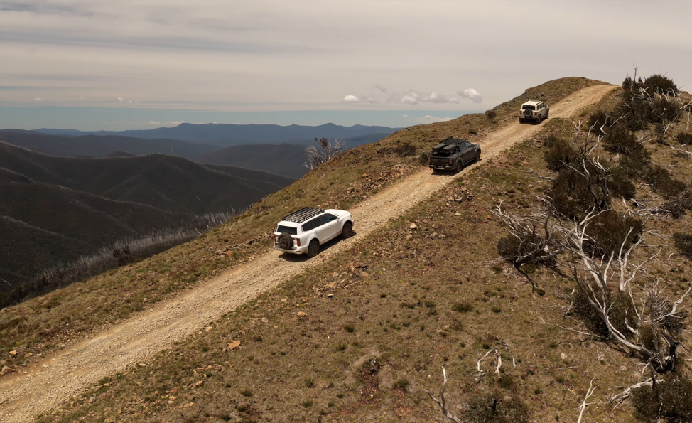 Three SUVs drive along a rugged mountain trail with a scenic view of distant hills under a cloudy sky. Sparse vegetation lines the path.