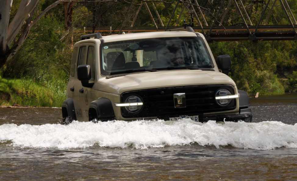 Four-wheel drive vehicle crossing a shallow river with a bridge and trees in the background.