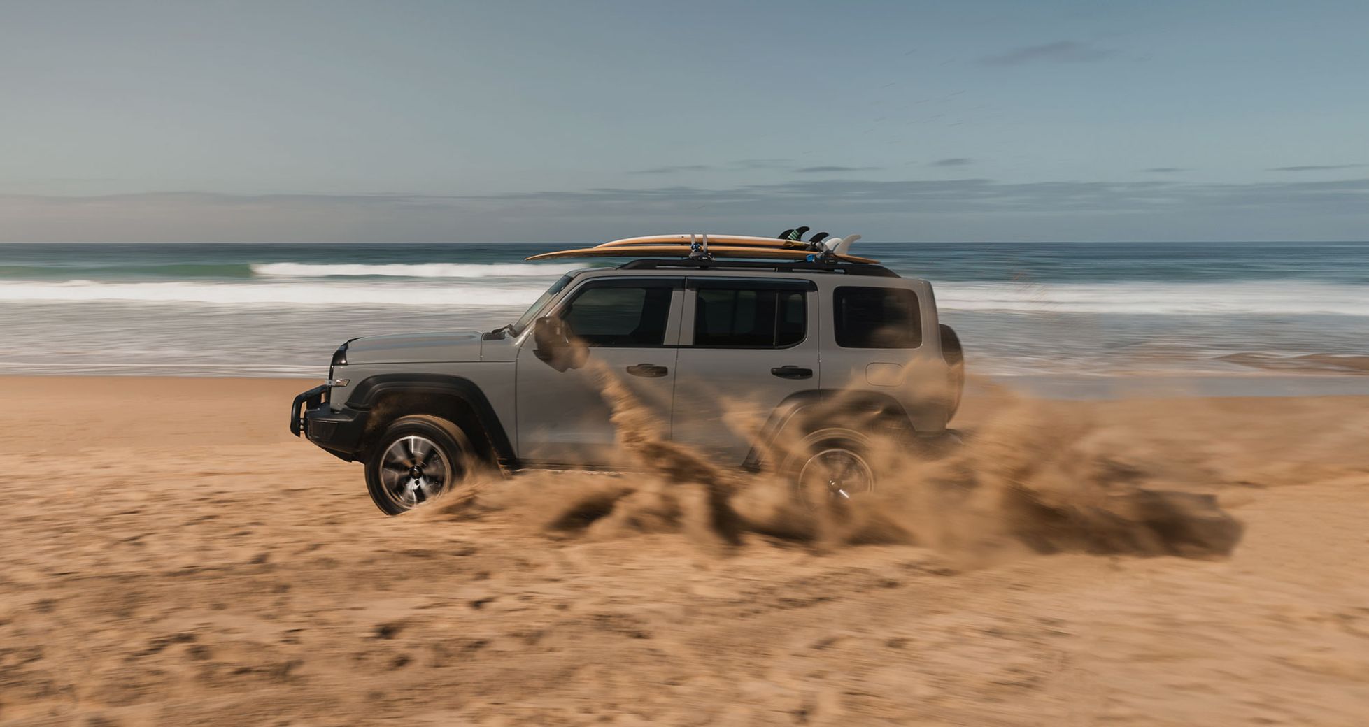Fossil Grey GWM Tank 300 with surfboards on the roof driving on the beach kicking up sand