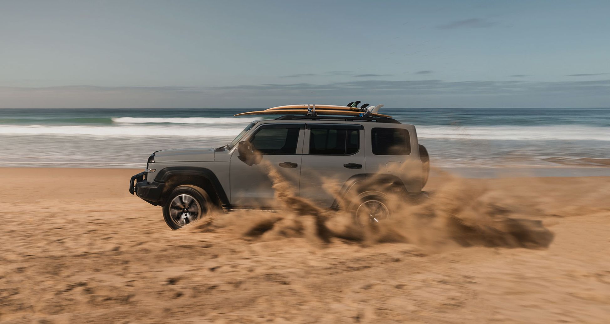 Fossil Grey GWM Tank 300 with surfboards on the roof driving on the beach kicking up sand