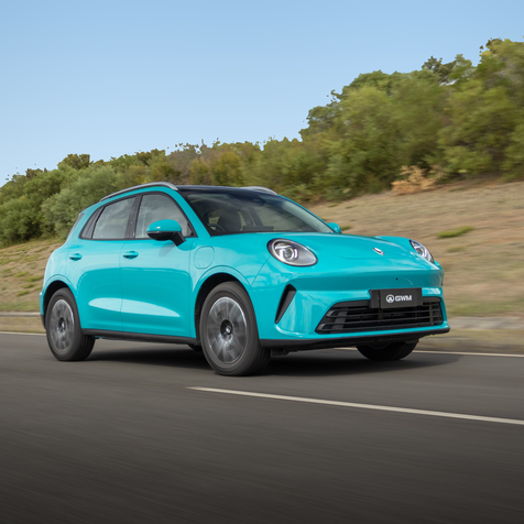 A blue hatchback car drives on a road with greenery in the background under a clear blue sky.