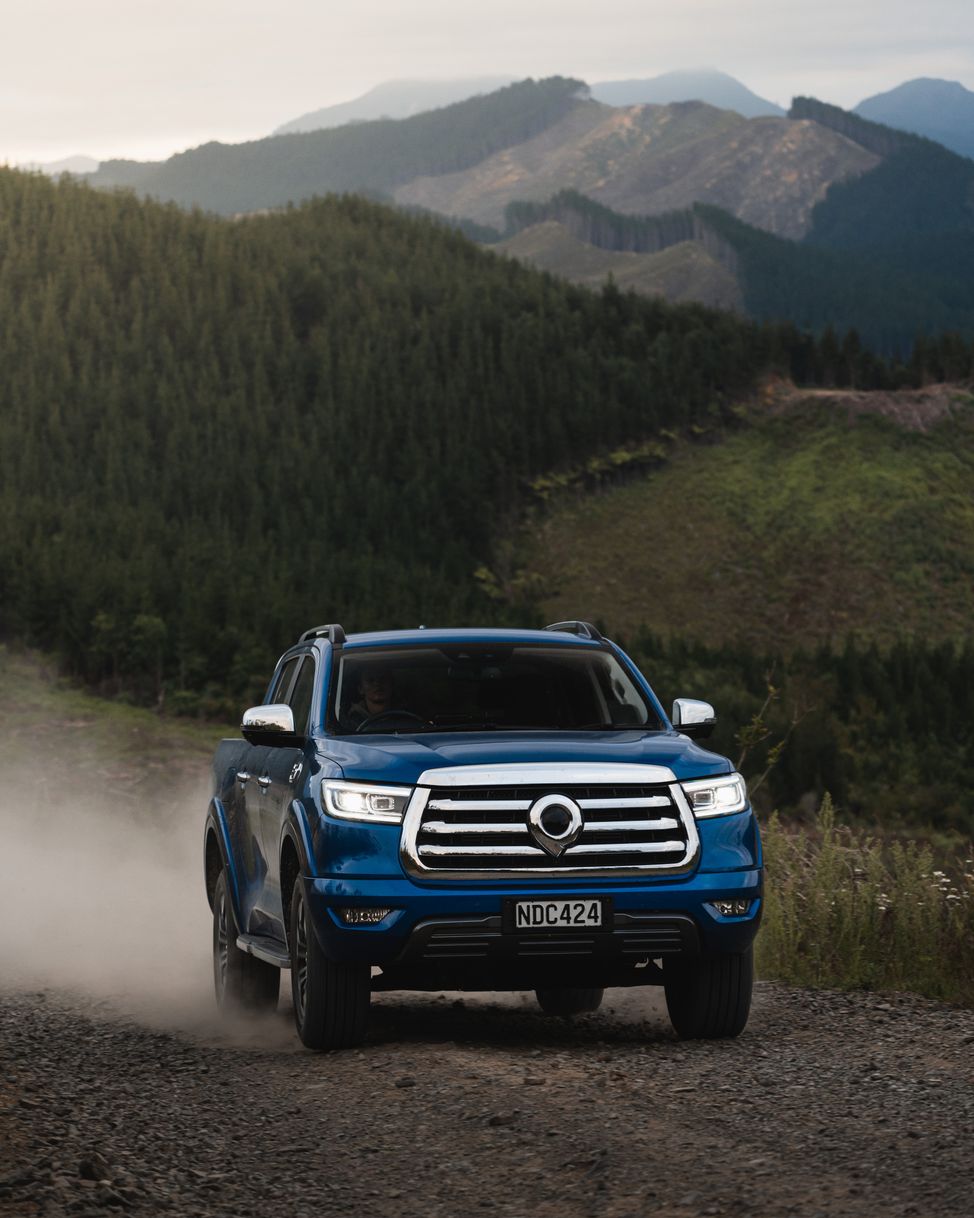 Sapphire Blue GWM Ute Cannon driving down a gravel road kicking up rocks and dust with mountains in the background