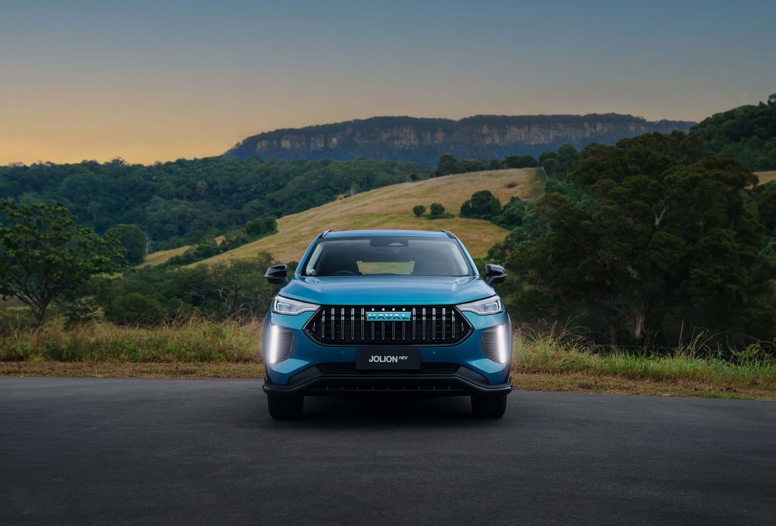 The front of GWM's small SUV, GWM Haval Jolion Hybrid in Aurora Green, parked in front of hills with trees with the foglights illuminated