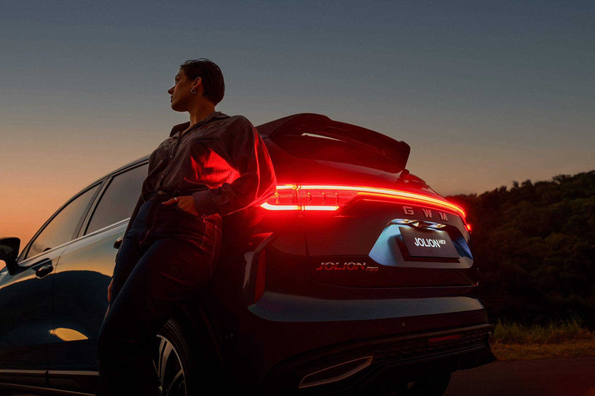 Female owner looking into the distance while leaning against GWM's small SUV - Aurora Green GWM Haval Jolion Hybrid as the sun is setting