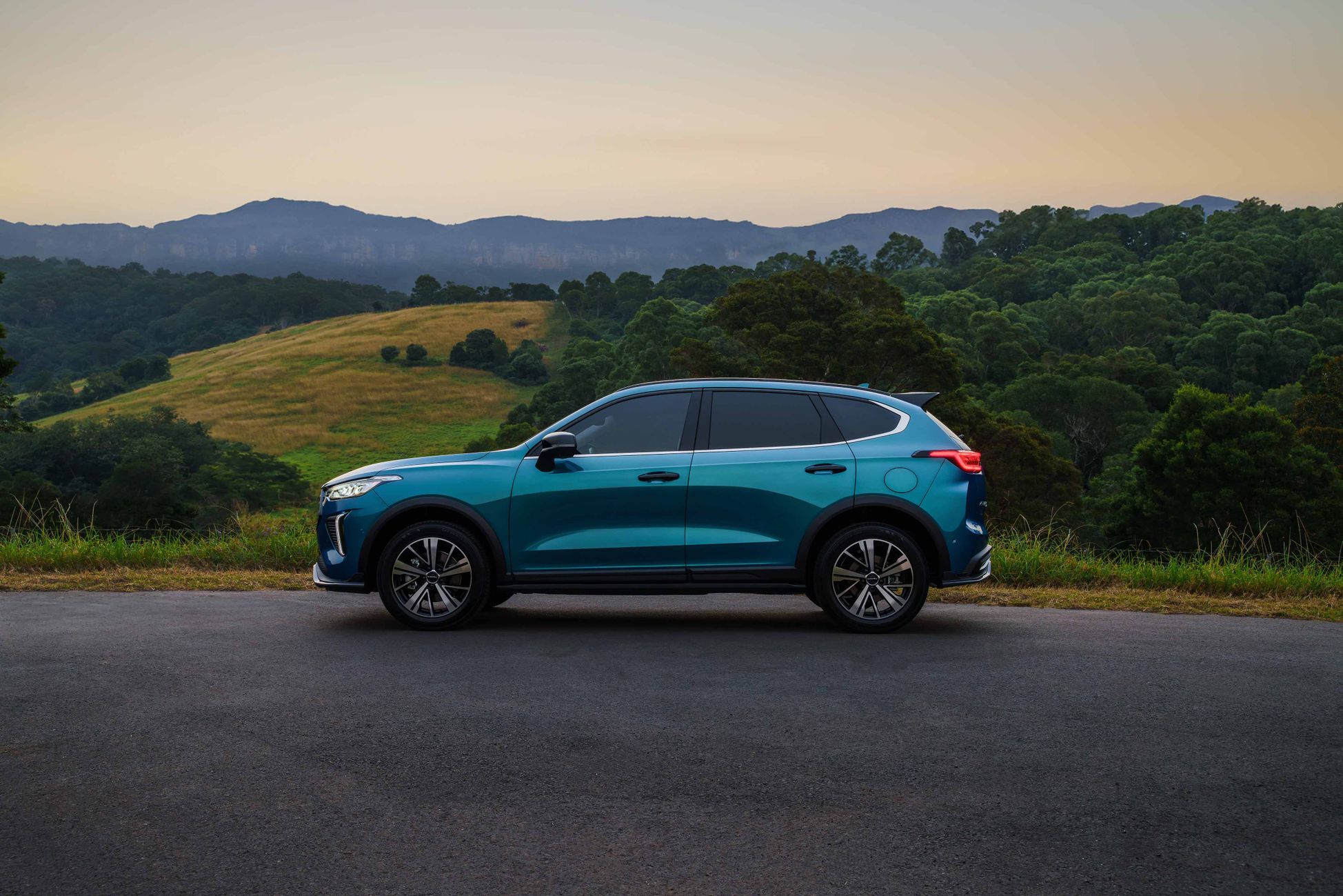 The side view of GWM's small SUV, GWM Haval Jolion Hybrid in Aurura Green, parked in front of a hill with trees