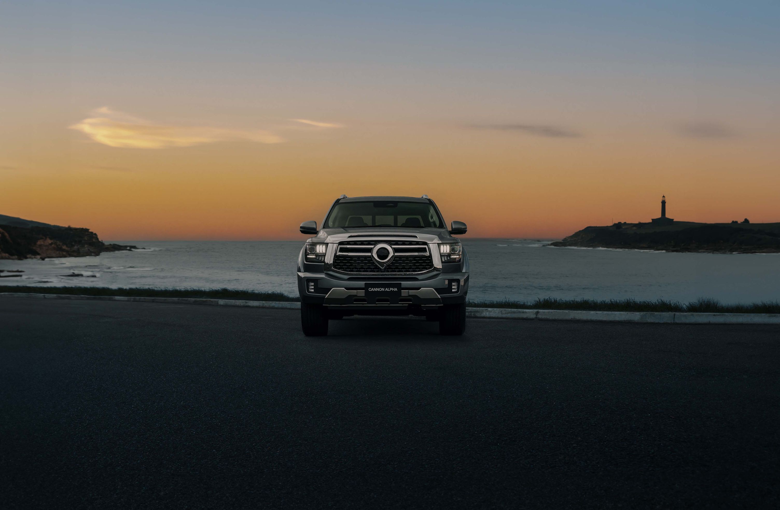 The front of a GWM Cannon Alpha in Onyx Silver, a luxury hybrid ute, parked on the beach with the sun setting behind the ocean