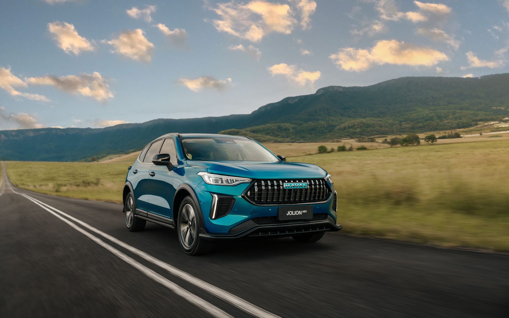 An Aurora Green GWM Haval Jolion Hybrid driving through the countryside with grass and mountains in the background