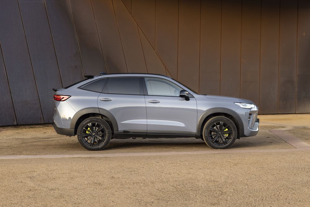 A Crayon Grey GWM Haval H6GT parked on yellow asphalt in front of a building with a steel exterior