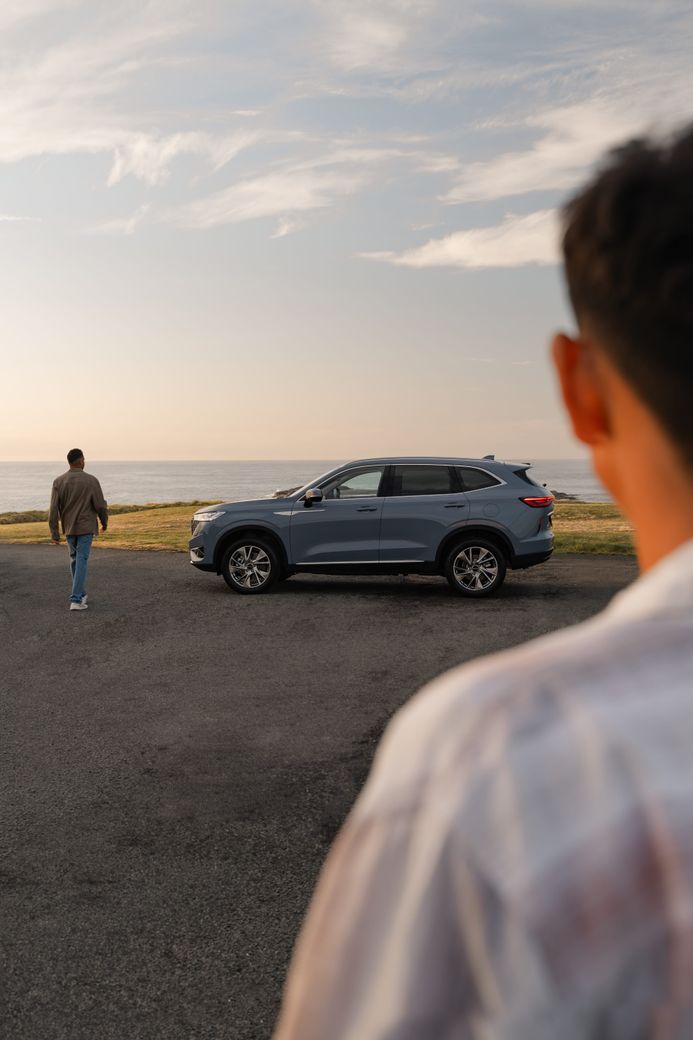 Two men walking towards a GWM Haval H6 Hybrid in Ayers Grey that is parked next to the sea on a gravel road