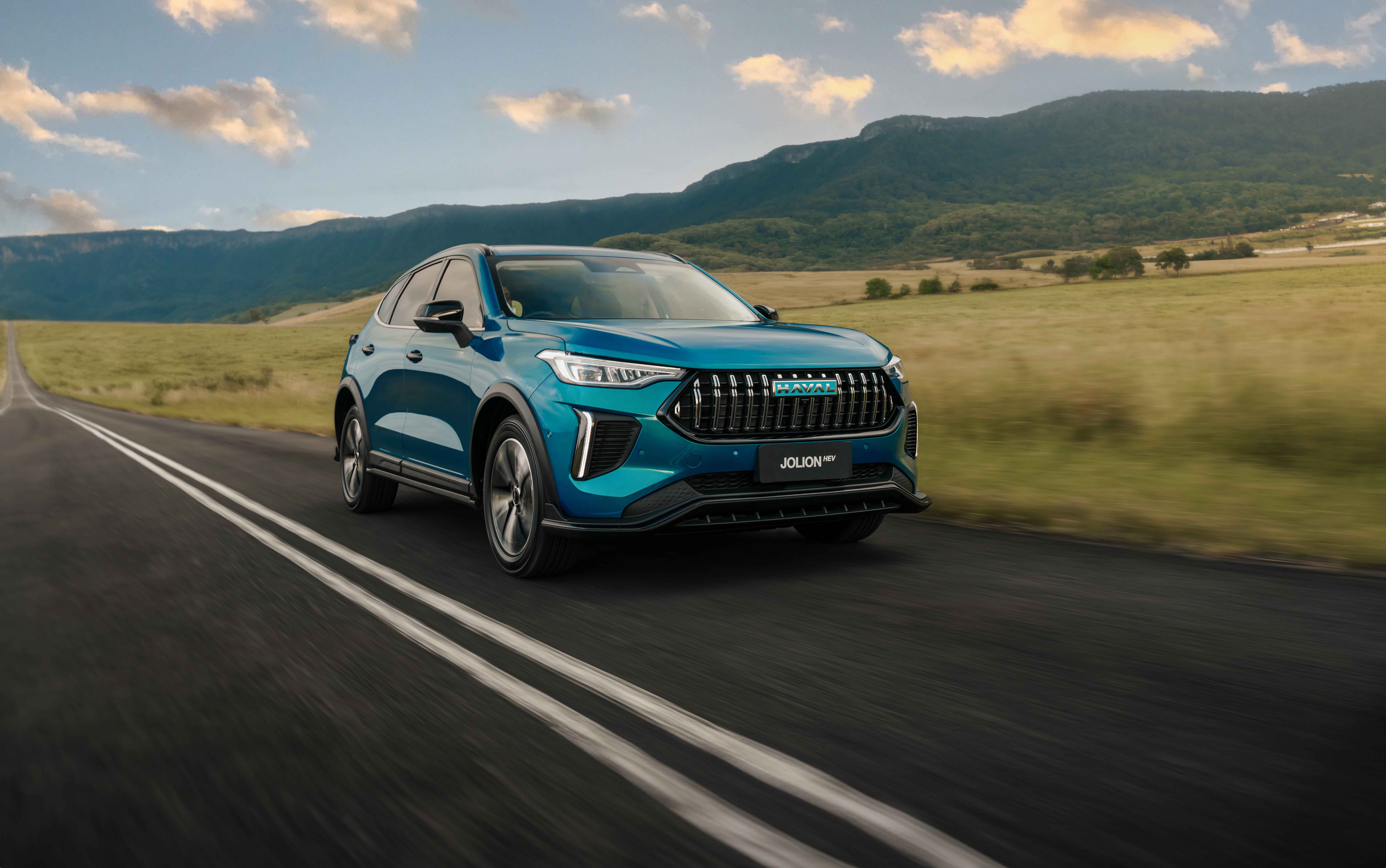 GWM Haval Jolion Hybrid in Aurora Green driving in the countryside with mountains in the background