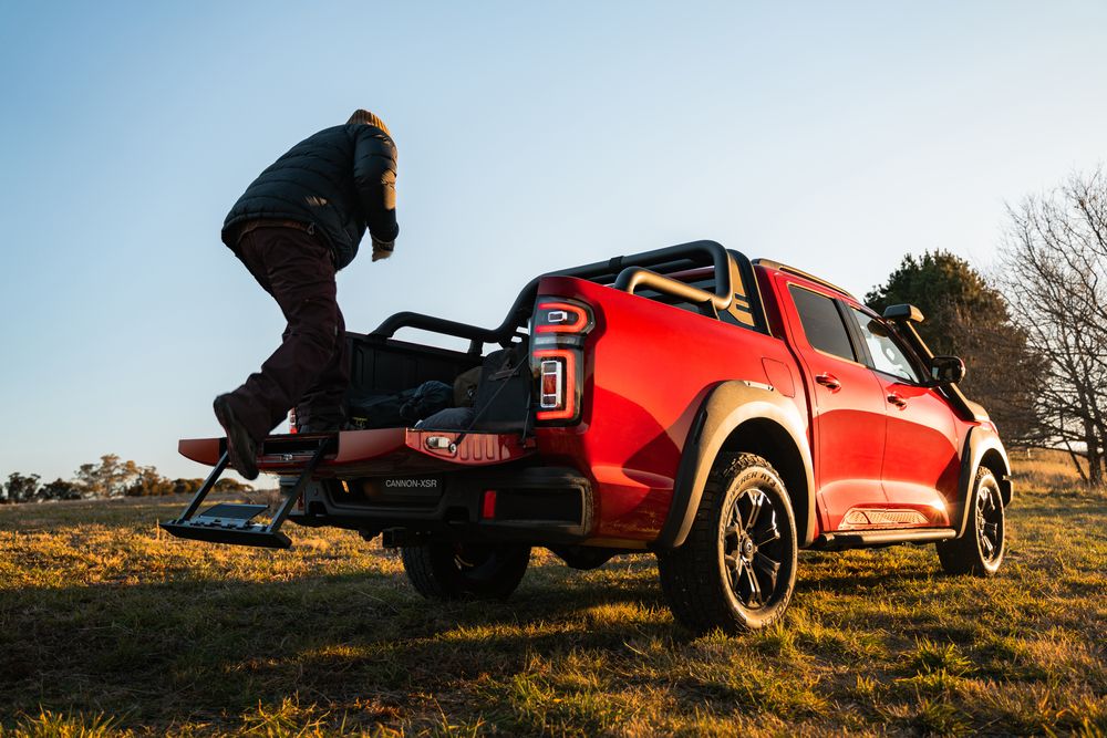 A Scarlet Red GWM Ute Cannon XSR parked on the grass in the middle of the wild with a man on the tailgate about to grab one of his bags stored in the ute tray
