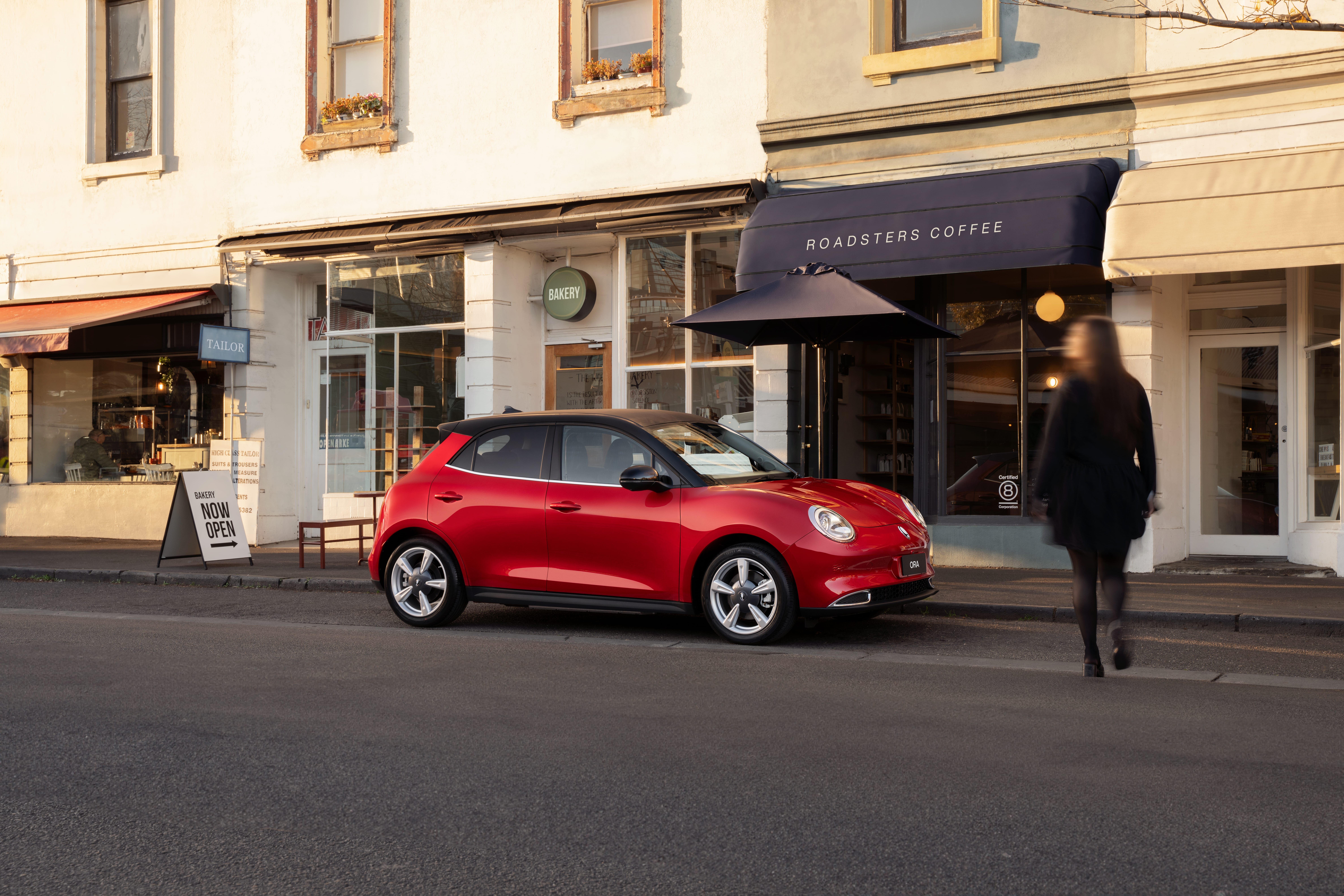 A Mars Red Ora with black roof parked in front of a city store front with a woman dressed in black walking towards it